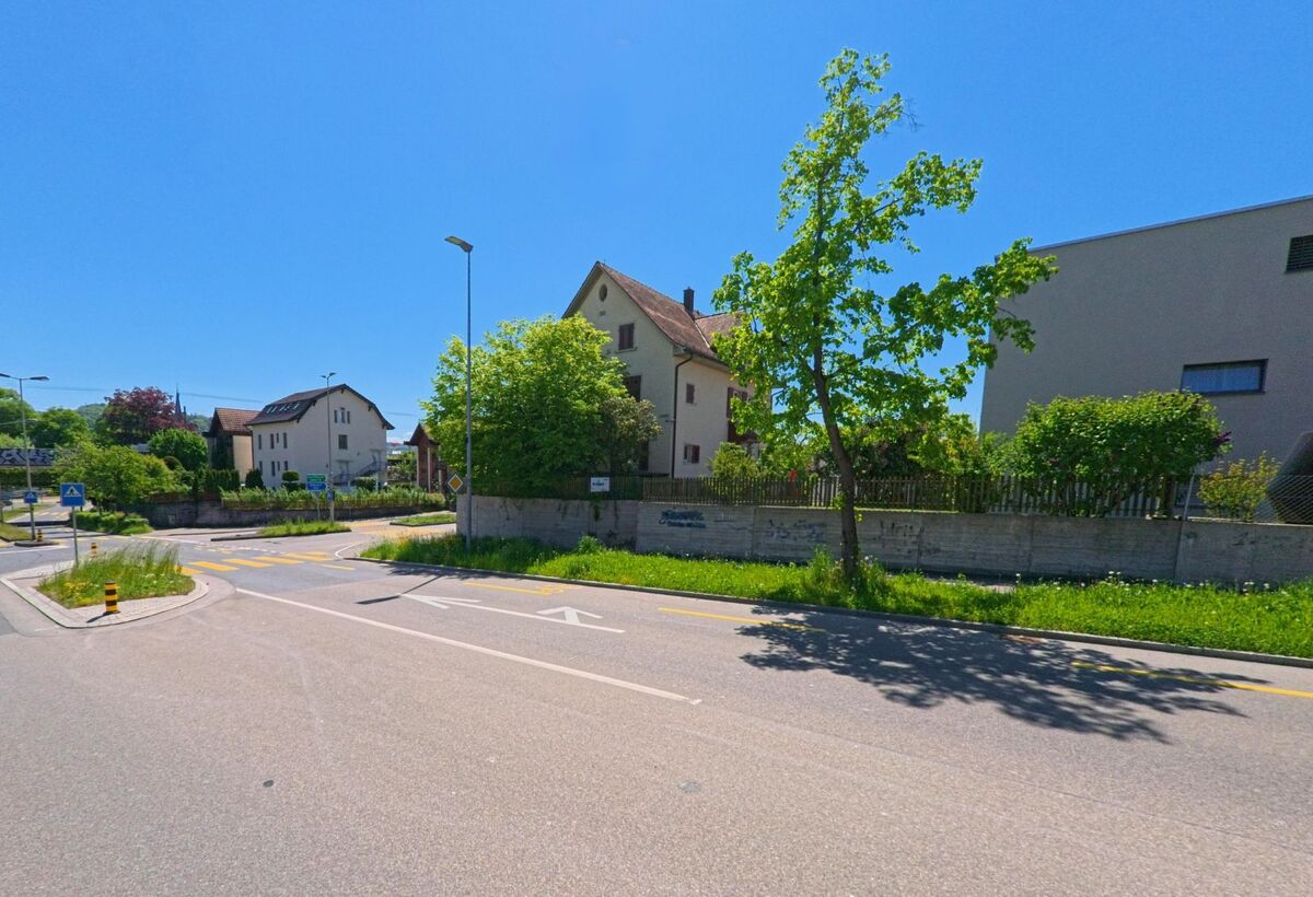 An einer Kreuzung in Bülach (8180, Kanton Zürich) stehen einzelne Wohnhäuser und ein Baum am Rand einer niedrigen Betonmauer bei strahlendem Sonnenschein.