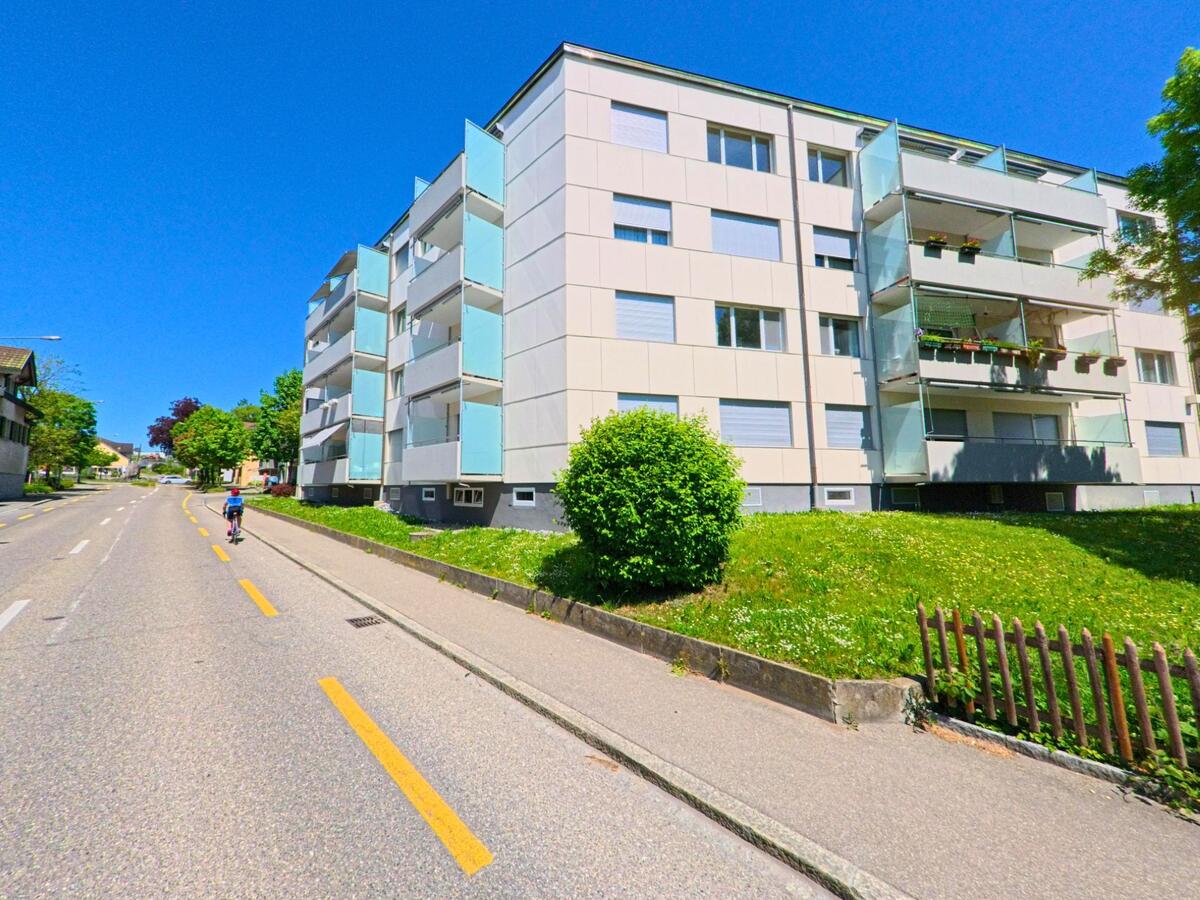 In Bülach im Kanton Zürich (PLZ 8180) säumen moderne Mehrfamilienhäuser mit Glasbalkonen eine Strasse mit separatem Radweg.
