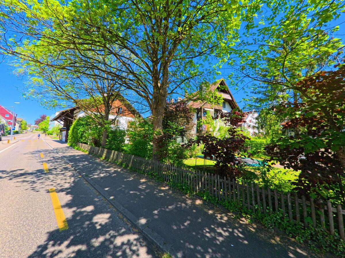In Bülach im Kanton Zürich (PLZ 8180) stehen traditionelle Wohnhäuser mit Gärten hinter einem alten Holzzaun entlang eines schattigen Rad- und Fusswegs.