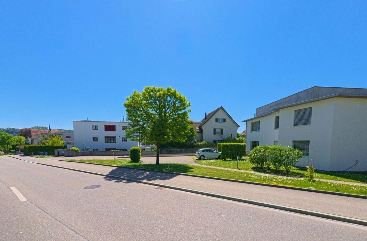  In Bülach (8180, Kanton Zürich) stehen moderne und ältere Wohnhäuser entlang einer ruhigen Strasse, gesäumt von gepflegtem Grün und einem grossen Baum im Vordergrund.