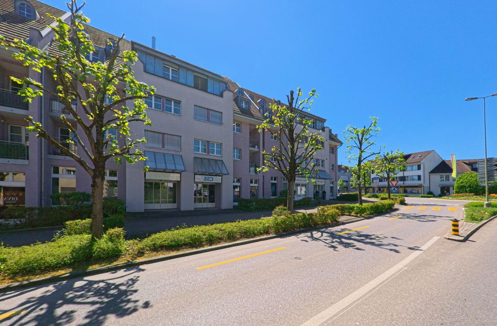 In Bülach (8180, Kanton Zürich) säumen frisch beschnittene Bäume eine ruhige Wohnstrasse mit mehrstöckigen Gebäuden und kleinen Geschäften im Erdgeschoss.