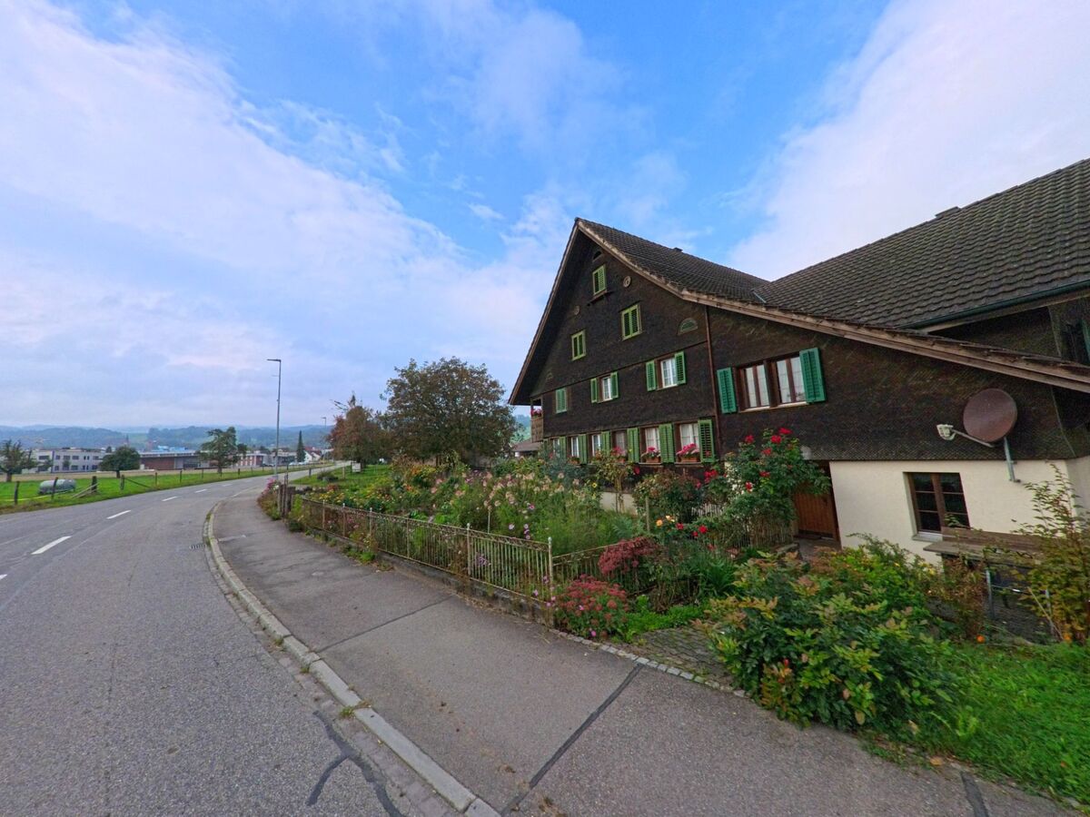 Ein traditionelles Bauernhaus mit dunkler Holzfassade und grünen Fensterläden steht in Bünzen (5624, Kanton Aargau) an einer leicht ansteigenden Strasse mit Blick auf das Tal.