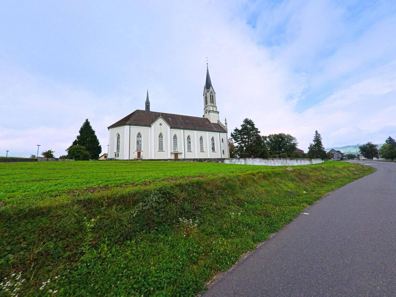 Die Kirche in Bünzen (5624, Kanton Aargau) steht leicht erhöht über einer grünen Wiese und ist von einem Weg mit Asphalt erreichbar.