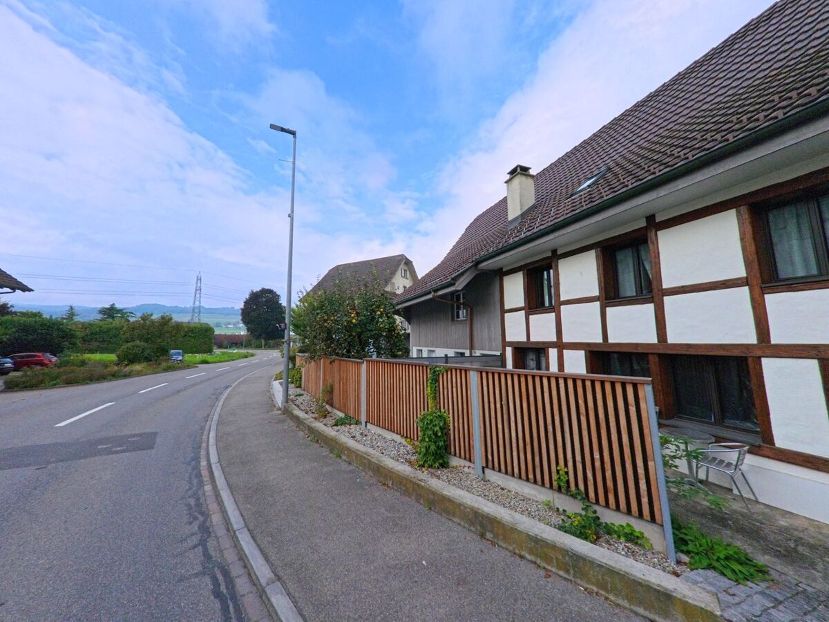 Ein traditionelles Fachwerkhaus mit braunen Holzbalken steht an einer leicht abfallenden Strasse in Bünzen (5624, Kanton Aargau) mit Blick auf das offene Landschaftsgebiet.