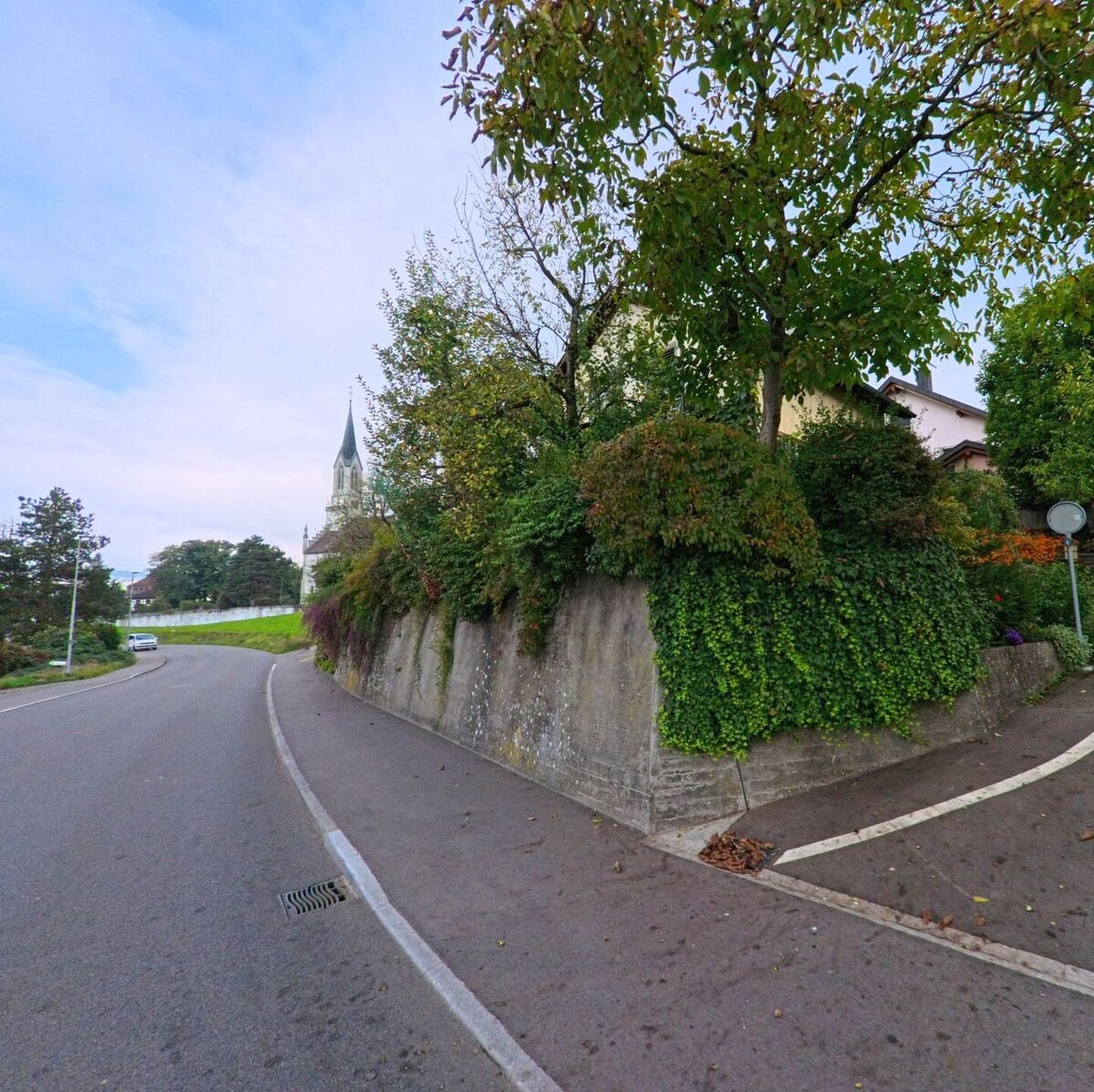 Eine ansteigende Strasse in Bünzen (5624, Kanton Aargau) führt entlang einer mit Efeu bewachsenen Mauer, im Hintergrund ist der Kirchturm der Dorfkirche sichtbar.