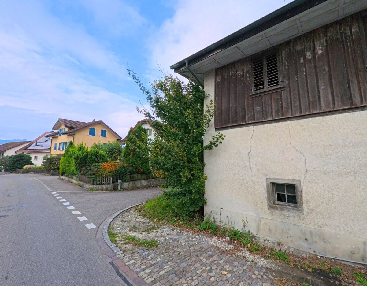 In einer Wohnstrasse von Bünzen (5624, Kanton Aargau) stehen ältere und neuere Häuser nebeneinander, gesäumt von Gärten und kleinen Vorgärten mit Sträuchern.