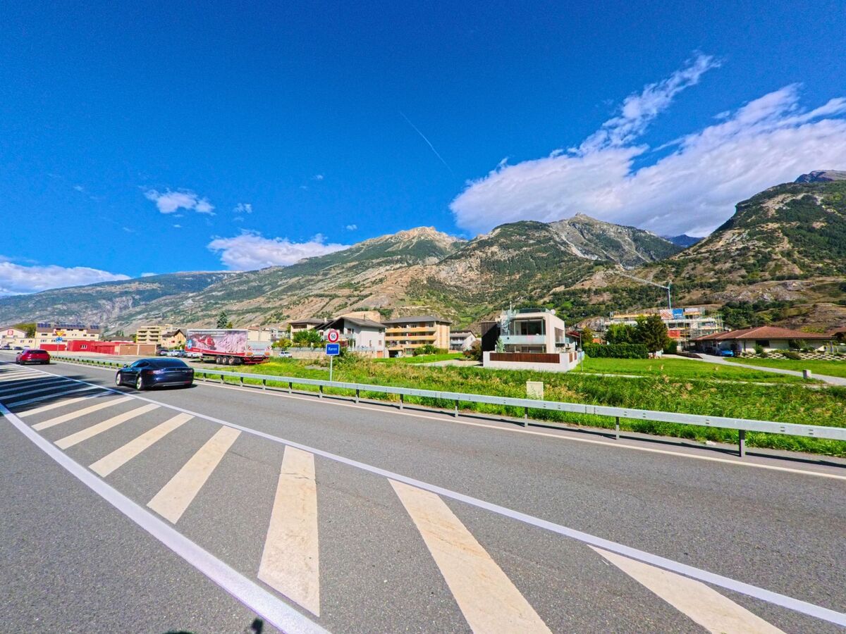 Autos fahren auf der Hauptstrasse unterhalb von Buerchen (3935, Kanton Wallis), im Hintergrund sind moderne Wohnhaeuser und die umliegenden Berge sichtbar.