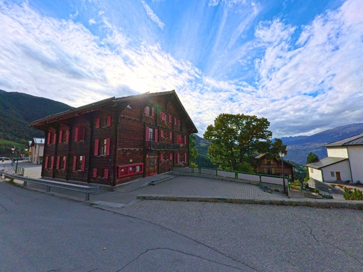 Ein traditionelles Holzhaus mit roten Fensterlaeden steht im Zentrum von Buerchen (3935, Kanton Wallis) vor einer Kulisse aus Bergen und bewaldeten Haengen.