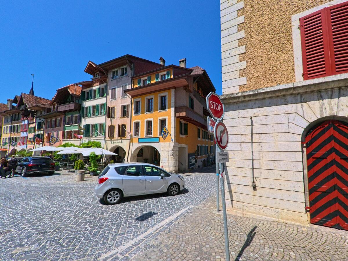 Eine gepflasterte Altstadtgasse in Büren an der Aare (3294, Kanton Bern) wird von farbigen Häusern mit Fensterlaeden und Balkonen gesaeumt, vor denen ein Strassencafé mit Sonnenschirmen eingerichtet ist.