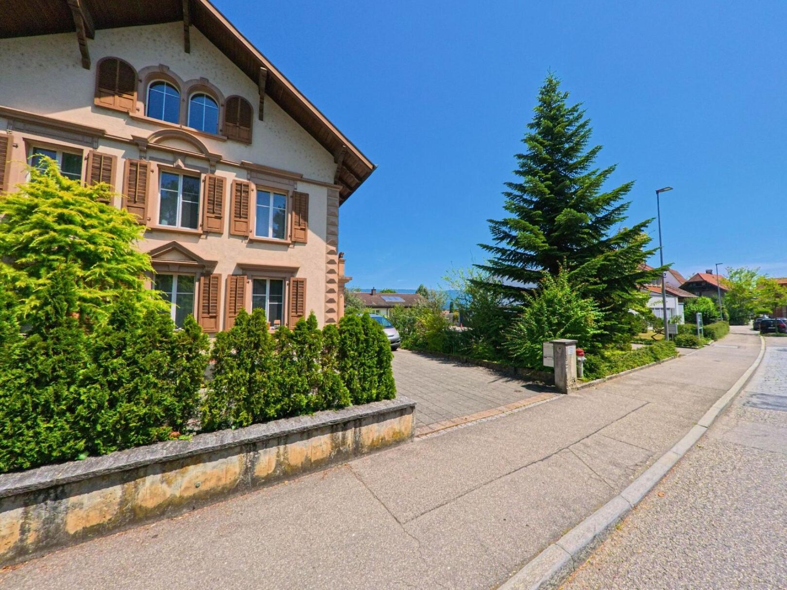 Eine gepflegte Quartierstrasse mit einem Wohnhaus in Büren an der Aare (3294, Kanton Bern) zeigt sich unter blauem Himmel und mit dichter Begrünung entlang des Gehwegs.