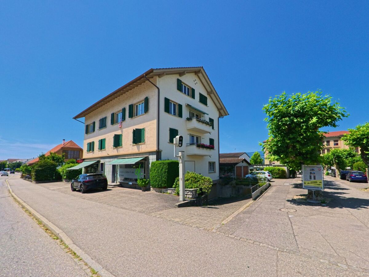 Ein Wohnhaus mit gruenen Fensterlaeden und einem Ladenlokal im Erdgeschoss steht in Büren an der Aare (3294, Kanton Bern) an einer ruhig verlaufenden Strasse mit Baumbepflanzung.