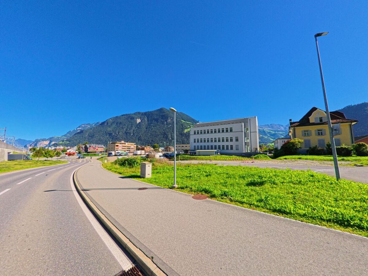 Eine breite Strasse in Bürglen (6463, Kanton Uri) fuehrt an modernen und historischen Gebaeuden vorbei, im Hintergrund erheben sich bewaldete Berge.