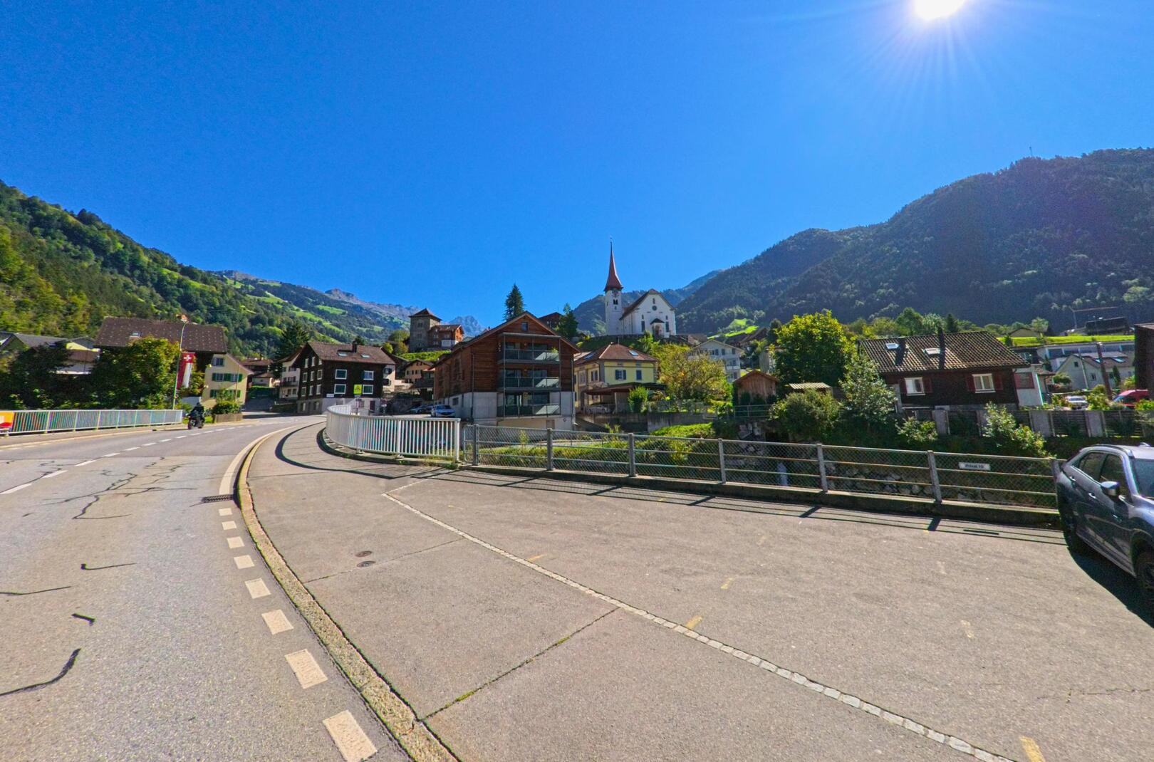 Bei klarem Himmel zeigt sich das Dorfzentrum von Bürglen (6463, Kanton Uri) mit der markanten Kirche und den umgebenden Bergen im Hintergrund.
