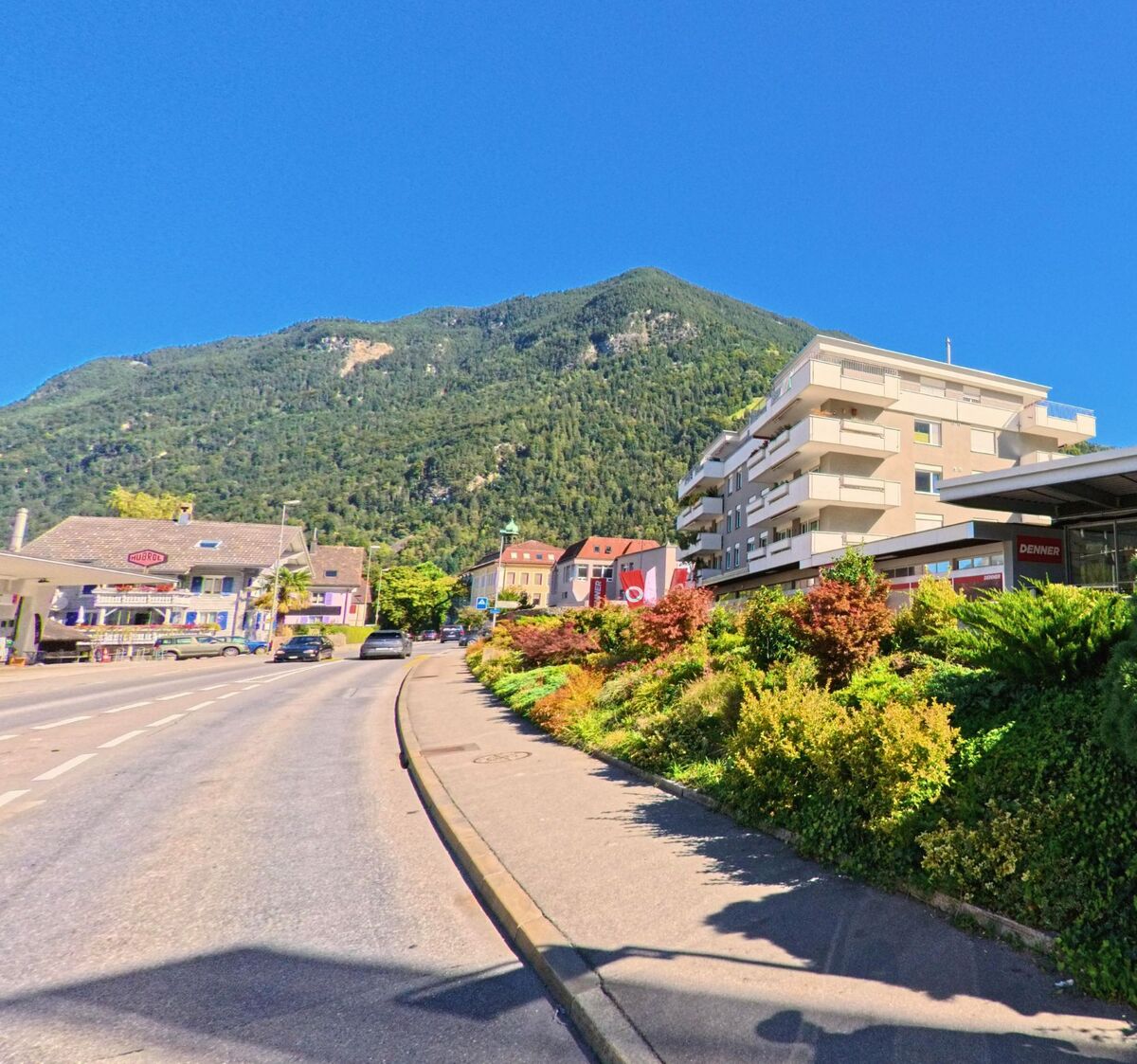 Entlang der Hauptstrasse von Bürglen (6463, Kanton Uri) stehen moderne und traditionelle Gebaeude, eingerahmt von bunten Pflanzen und dem bewaldeten Berg im Hintergrund.