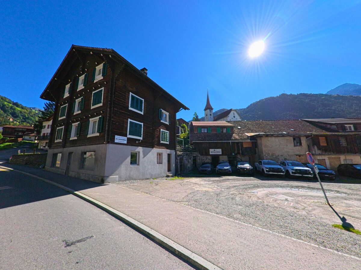 Ein traditionelles Holzhaus in Bürglen (6463, Kanton Uri) steht an einer sonnigen Strasse, im Hintergrund ist die Kirche mit ihrem markanten Turm zu sehen.