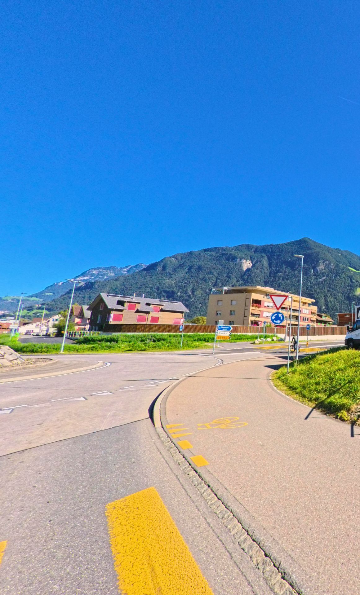 Ein Kreisverkehr in Bürglen (6463, Kanton Uri) liegt vor moderner Wohnbebauung und gruenen Berghaengen unter strahlend blauem Himmel.