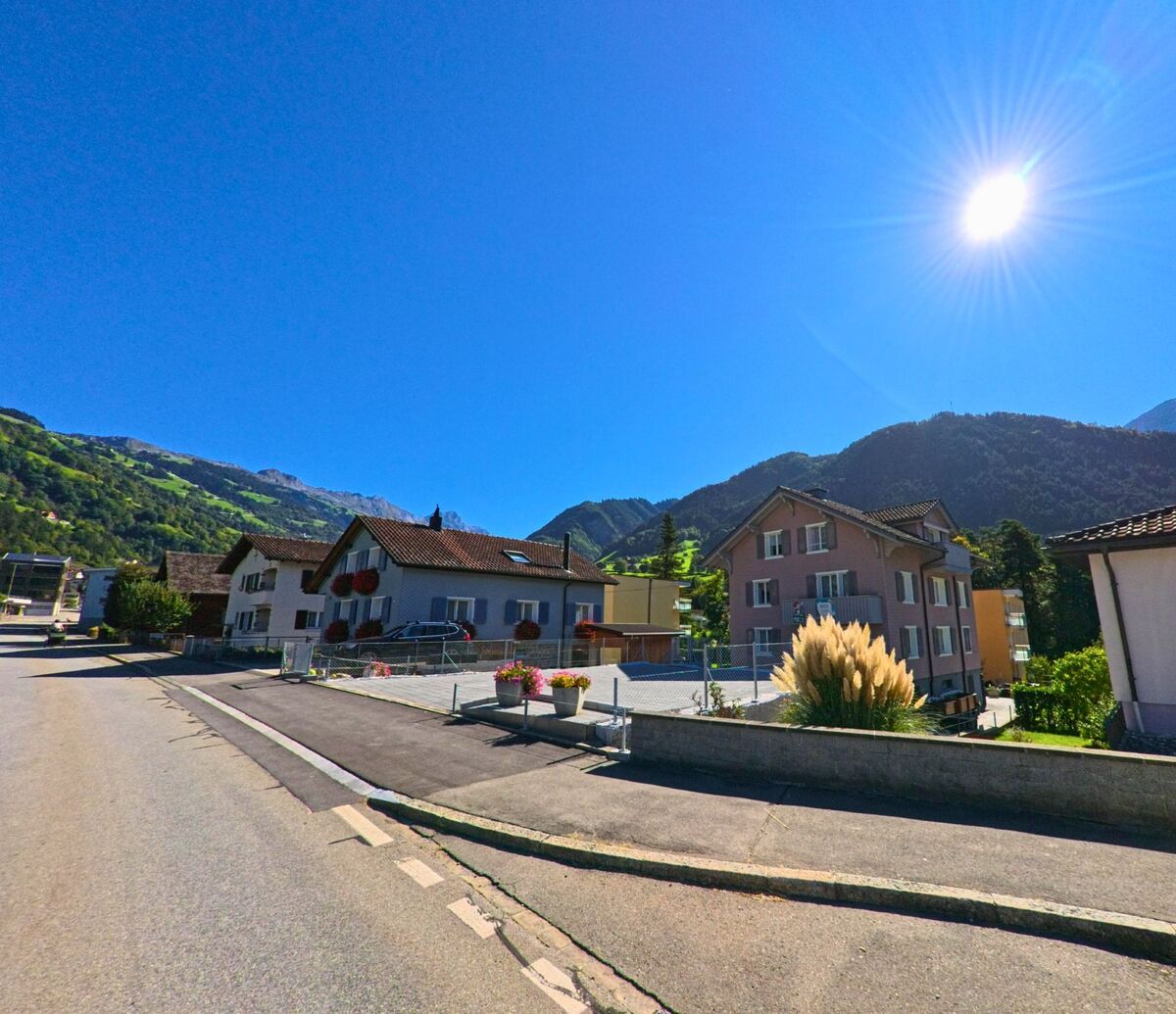Unter strahlender Sonne liegen mehrere Wohnhaeuser in Bürglen (6463, Kanton Uri) am Hang mit Blick auf die umliegenden gruenen Berge.