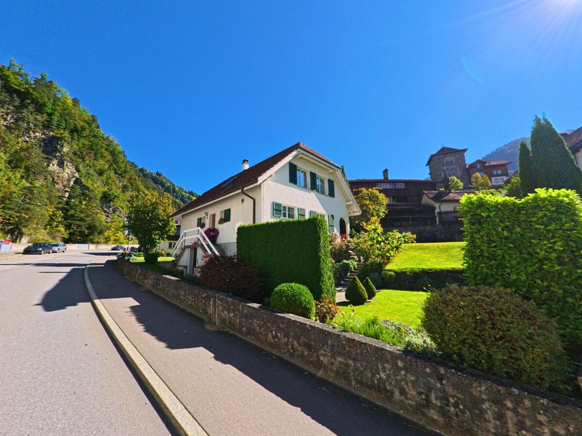 Ein gepflegtes Wohnhaus mit gruenem Garten in Bürglen (6463, Kanton Uri) liegt an einer leicht ansteigenden Strasse vor bewaldeten Haengen und alten Gebaeuden.