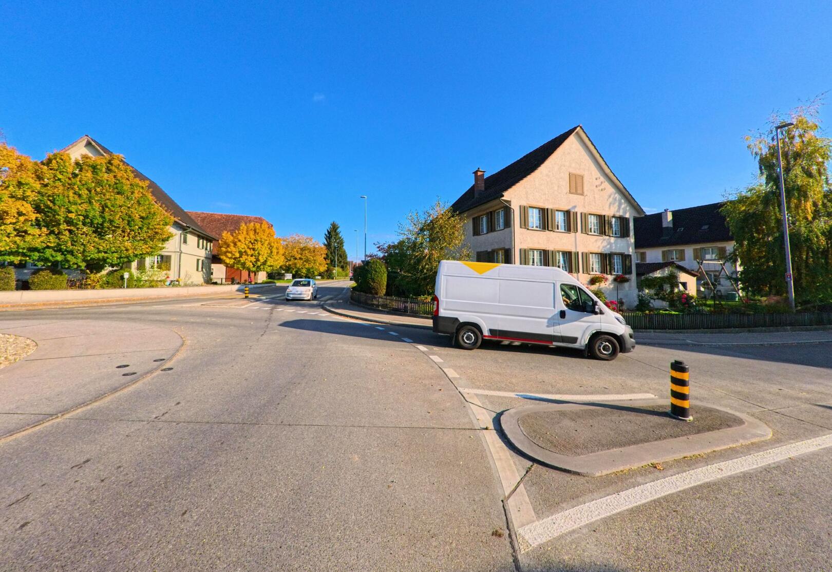 An einer breiten Kreuzung in Bürglen (8575, Kanton Thurgau) biegt ein weisser Lieferwagen bei Herbstsonne an Wohnhäusern und bunten Bäumen vorbei ab.