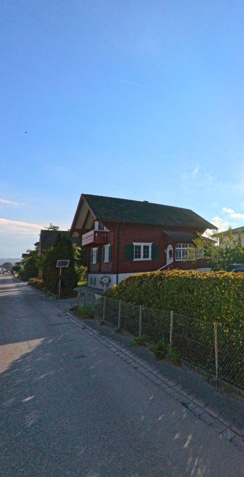 An einer schmalen Quartierstrasse in Bürglen (8575, Kanton Thurgau) steht ein rotes Haus mit grünen Fensterläden hinter einer niedrigen Hecke im Abendlicht.