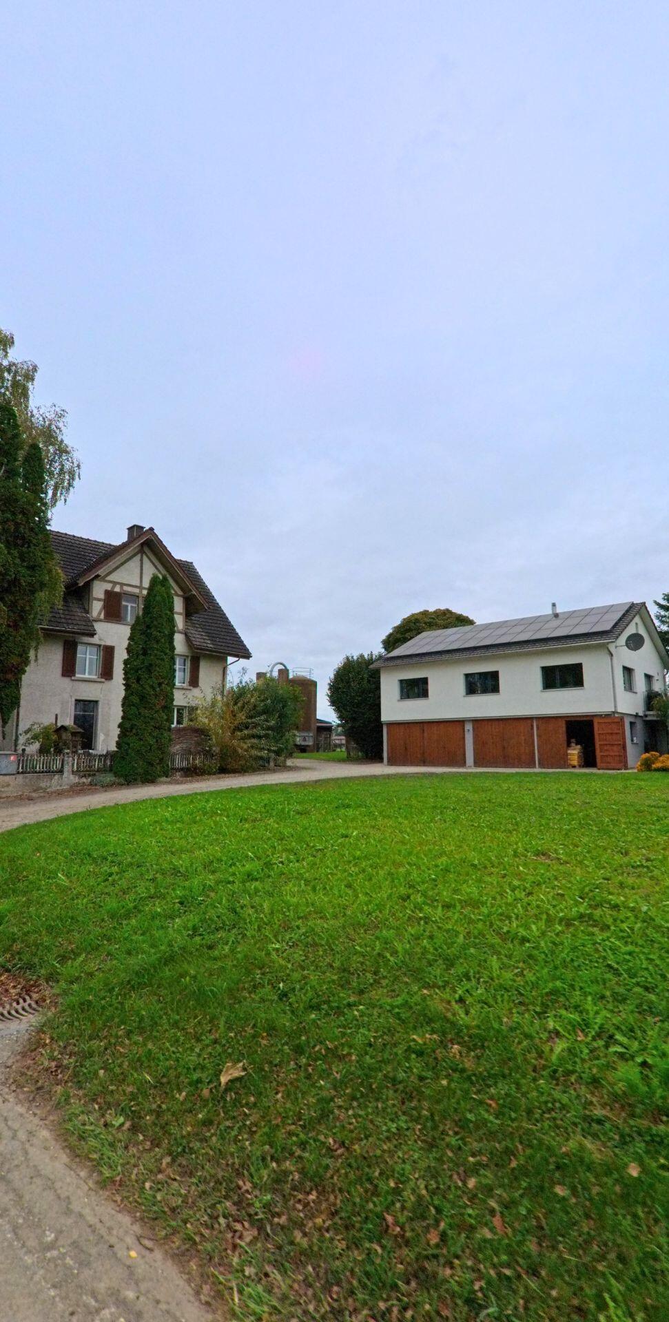 Neben einer grossen Wiese in Bürglen (8575, Kanton Thurgau) stehen ein älteres Wohnhaus und ein Wirtschaftsgebäude mit Holztoren und Solarpanels auf dem Dach.