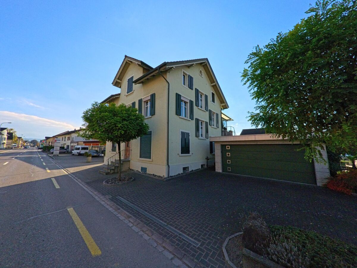 Am Strassenrand in Bürglen (8575, Kanton Thurgau) steht ein helles Wohnhaus mit grünen Fensterläden neben einer breiten Garageneinfahrt und Bäumen.