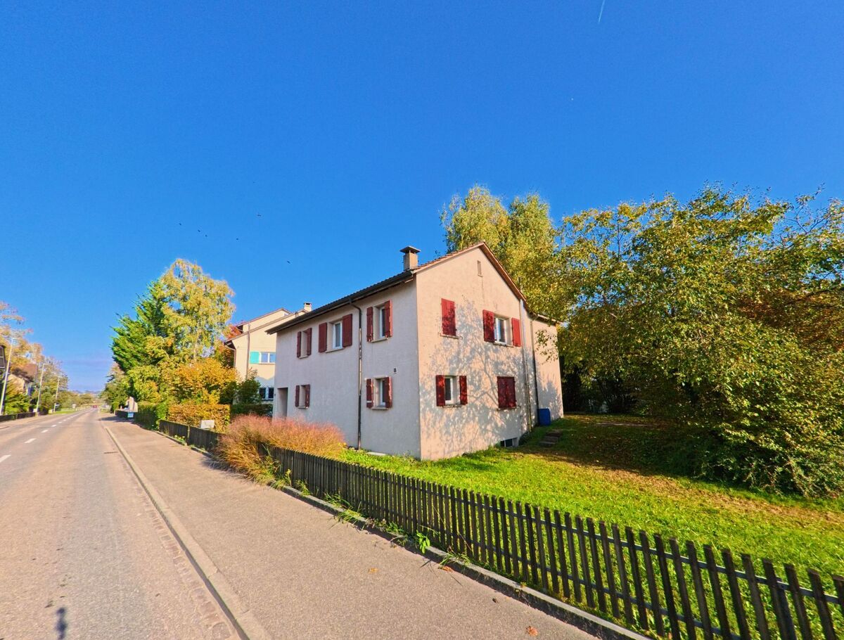 Ein helles Wohnhaus mit roten Fensterläden steht in Bürglen (8575, Kanton Thurgau) neben einer Strasse, umgeben von Gartenfläche und Bäumen.
