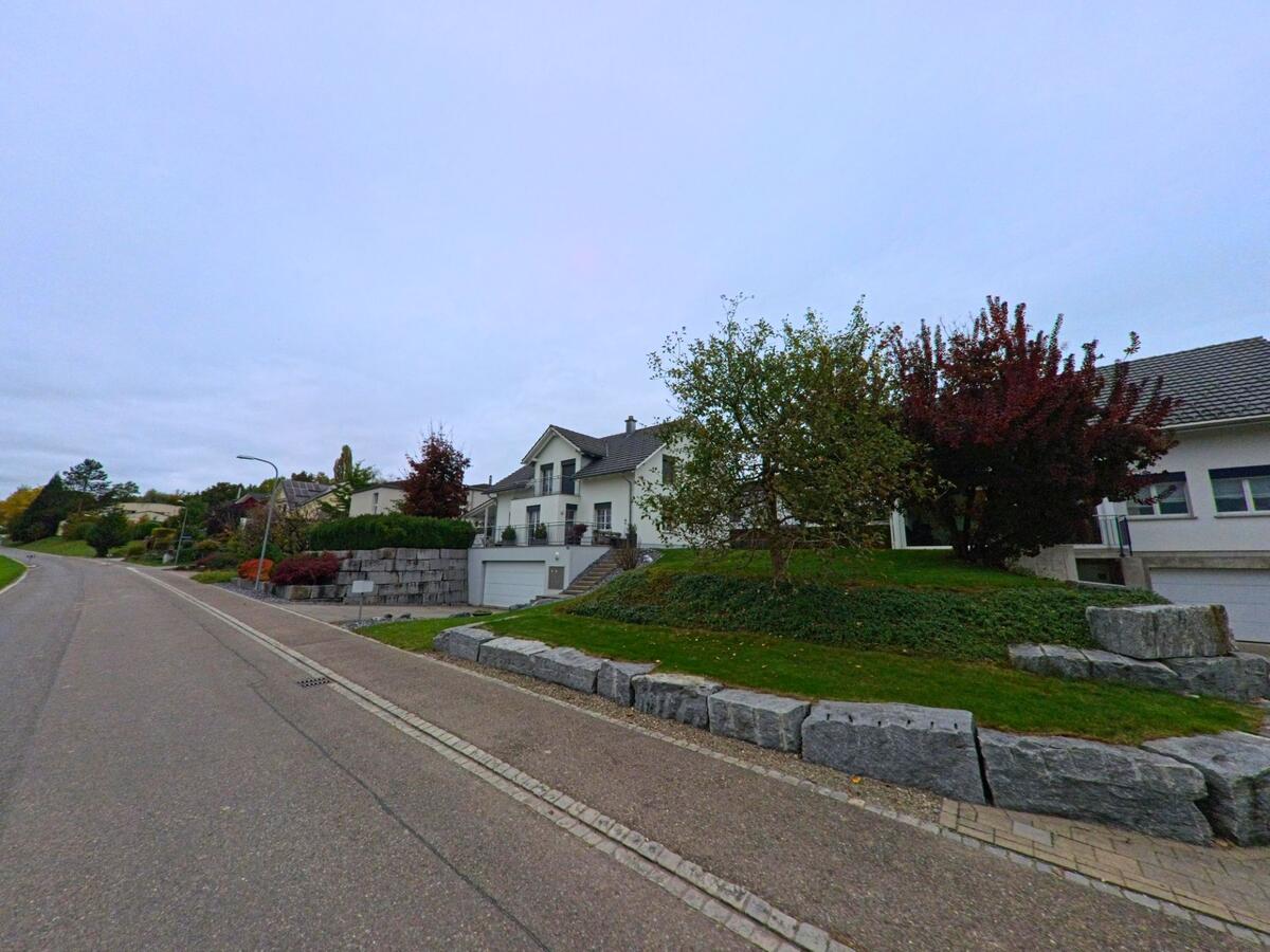 Eine leicht ansteigende Wohnstrasse in Bürglen (8575, Kanton Thurgau) führt an gepflegten Gärten, Steinmauern und Einfamilienhäusern vorbei.