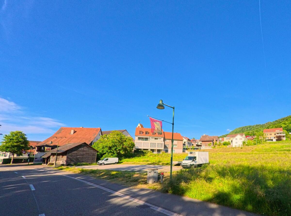 Vom Randbereich in Büsserach (4227, Kanton Solothurn) fällt der Blick über eine freie Wiese hinweg auf das Dorfzentrum mit Kirche, Wohnhäusern und Fahnenmasten.