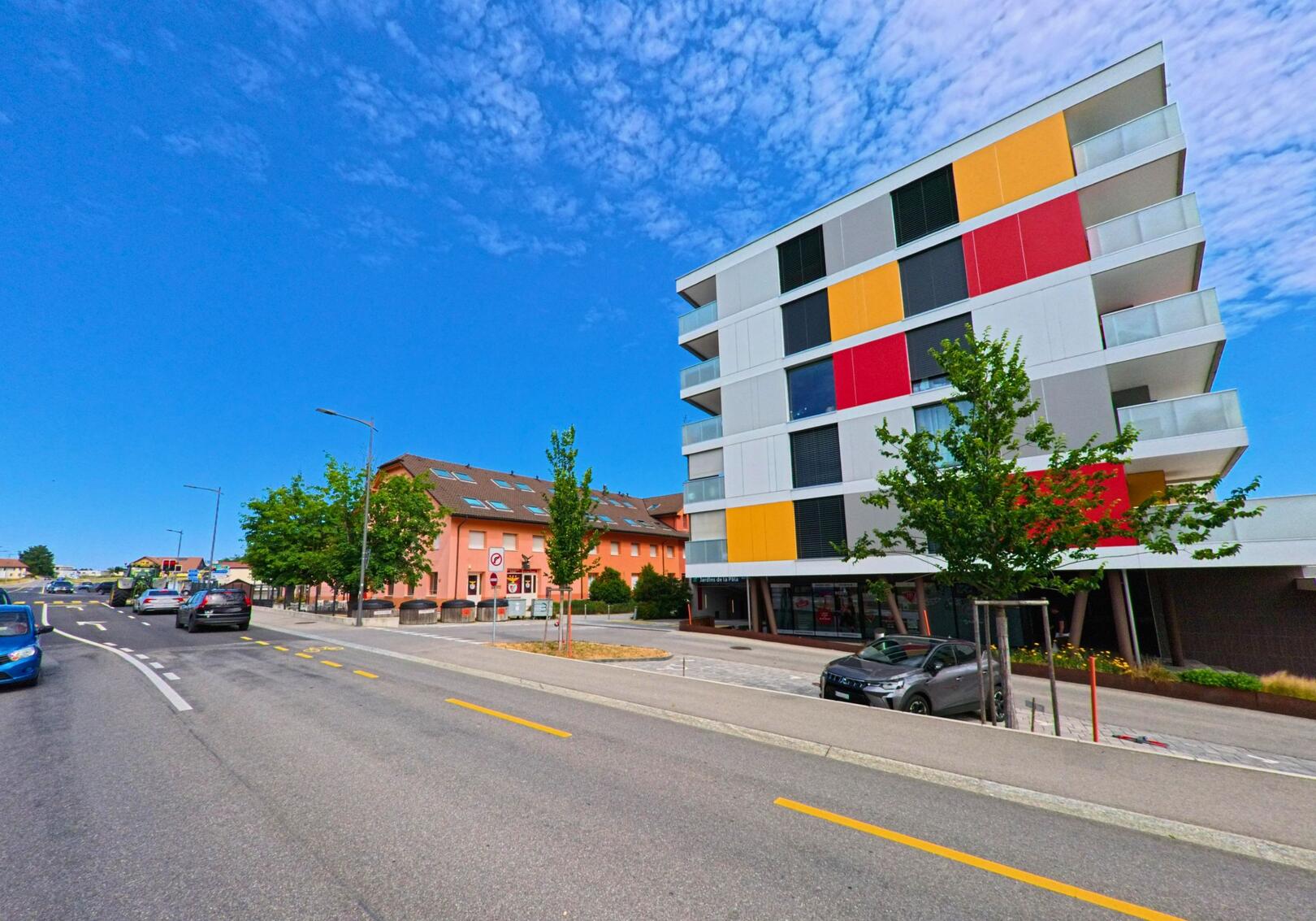 In Bulle (1630, Kanton Freiburg) stehen moderne Wohngebäude mit farbigen Fassadenelementen entlang einer verkehrsreichen Strasse mit separatem Velostreifen.