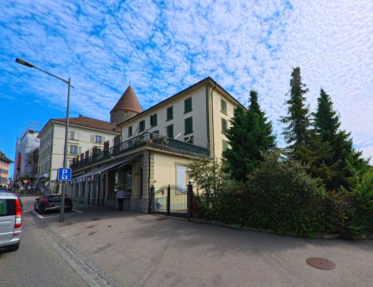 In Bulle (1630, Kanton Freiburg) ragt ein mittelalterlicher Rundturm hinter historischen Gebäuden und dichtem Grün hervor, flankiert von einem Gehweg mit Parkplätzen.