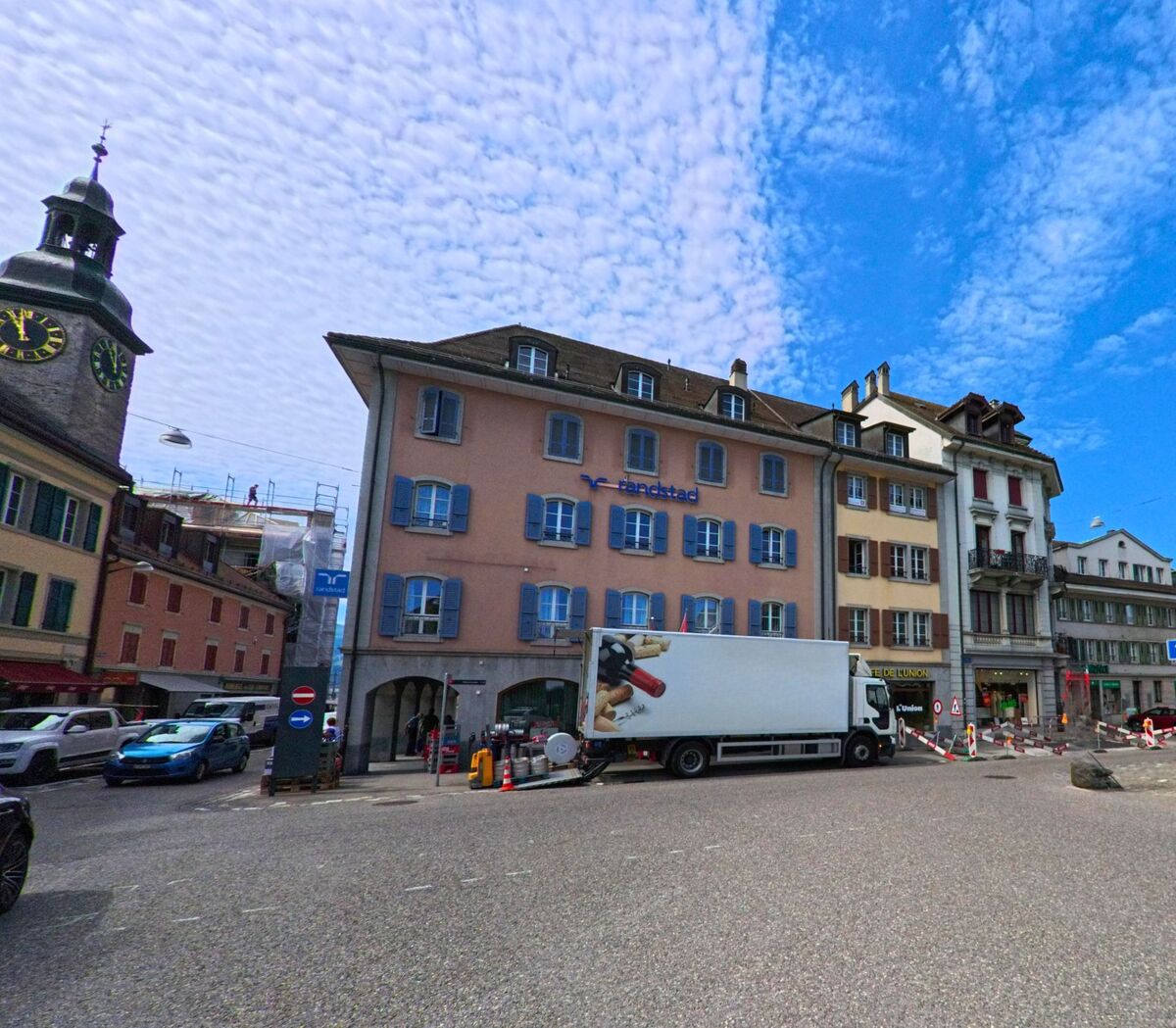 In der Altstadt von Bulle (1630, Kanton Freiburg) stehen farbenfrohe Gebäude mit blauen Fensterläden an einer belebten Kreuzung, neben einem markanten Uhrturm mit Zwiebelhaube.