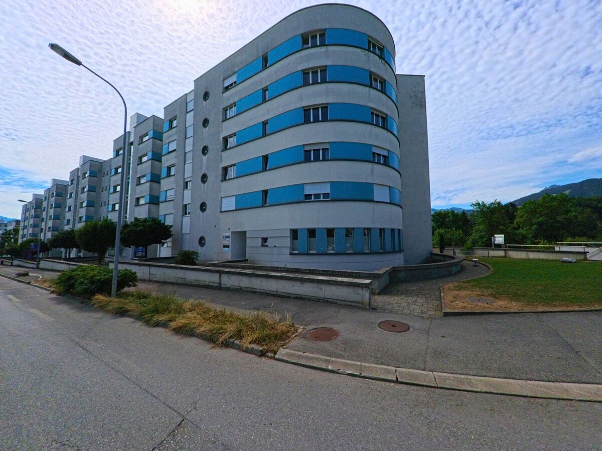 Ein langgestreckter Wohnblock mit abgerundeter Ecke und blauen Fensterbändern steht in Bulle (1630, Kanton Freiburg) an einer ruhigen Seitenstrasse mit Blick ins Grüne.