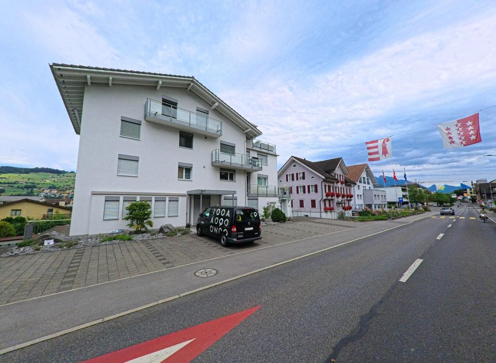 Entlang der Hauptstrasse in Buochs (6374, Kanton Nidwalden) stehen moderne und traditionelle Wohnhäuser, geschmückt mit Fahnen verschiedener Schweizer Kantone.