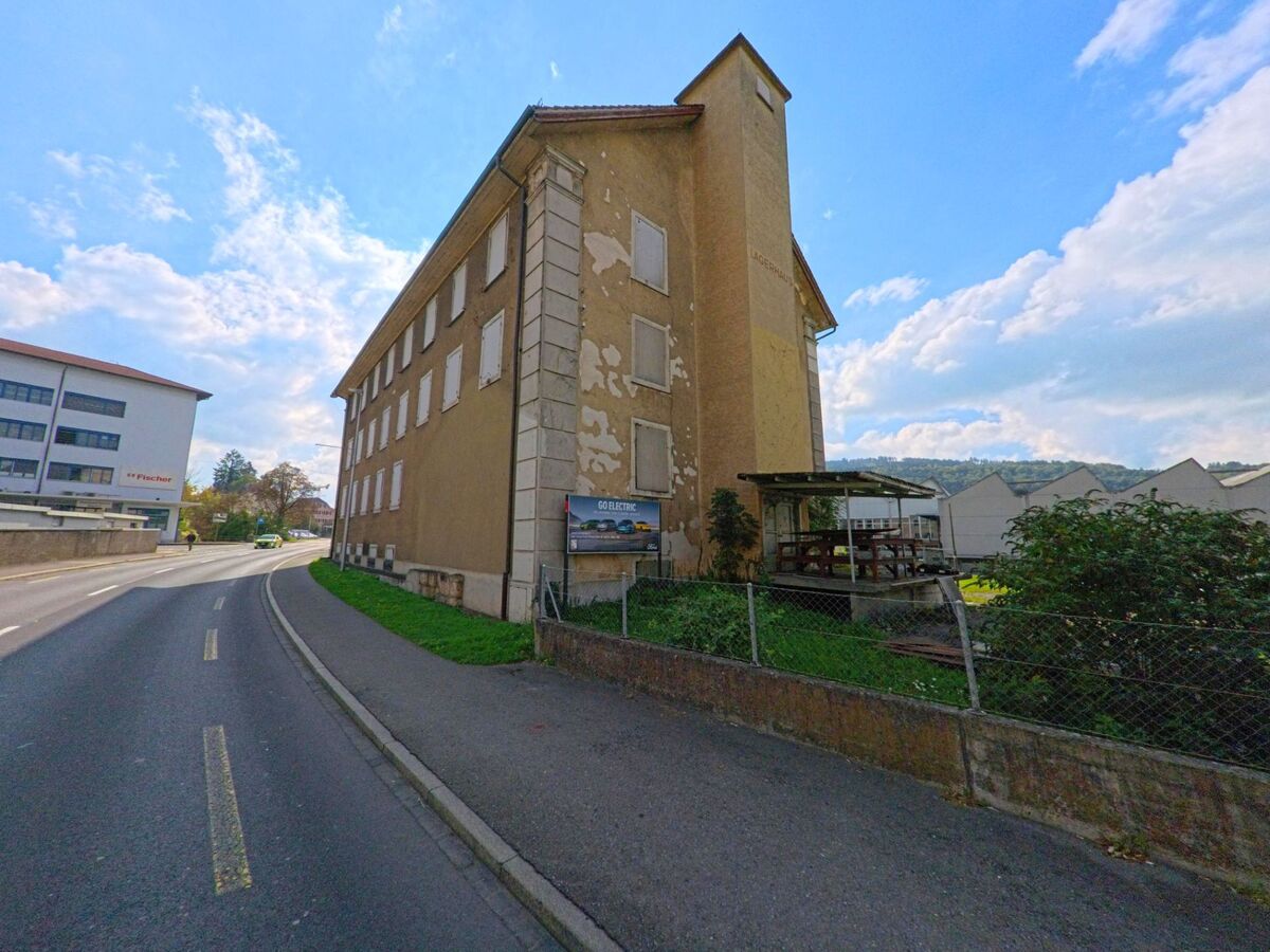Ein altes, mehrstöckiges Lagerhaus mit verblasster Fassade steht an der Hauptstrasse von Burg (5734, Kanton Aargau) neben modernen Gewerbebauten und Gewächshäusern.