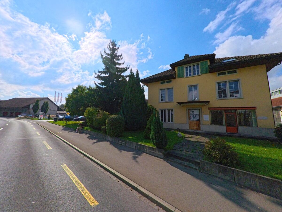 An der Hauptstrasse von Burg (5734, Kanton Aargau) steht ein gelbes Wohnhaus mit grünen Fensterläden, umgeben von gepflegtem Rasen und Bäumen unter blauem Himmel.