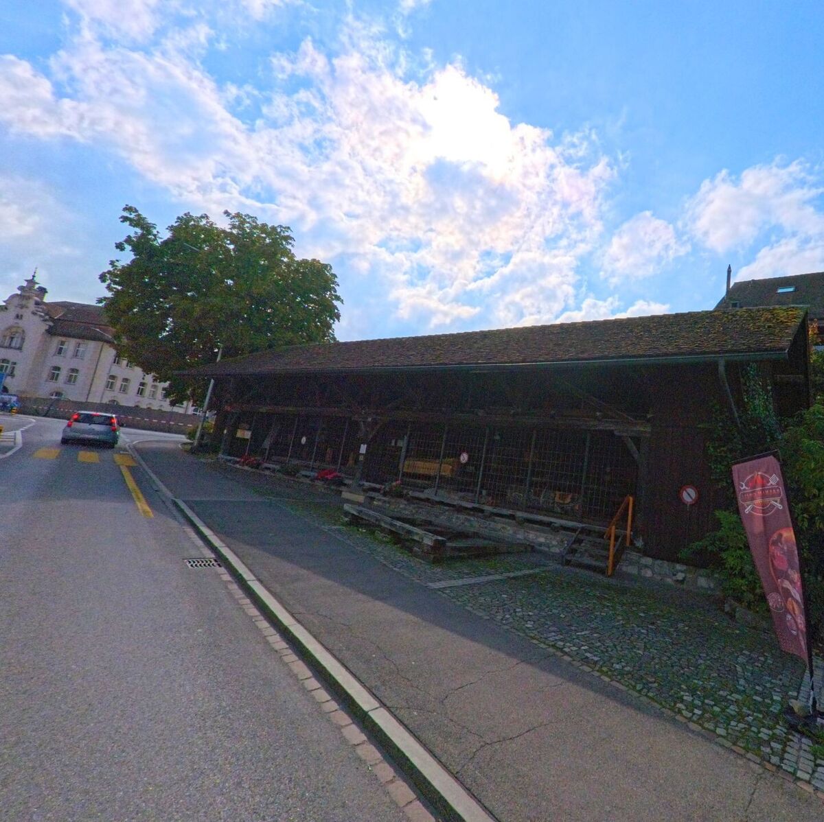Im Zentrum von Burg (5736, Kanton Aargau) steht ein langes Holzgebäude mit überdachter Fassade an einer leicht gekrümmten Strasse nahe einem historischen Haus.