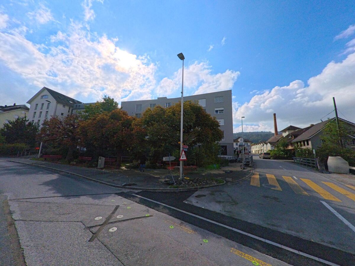 An einer Kreuzung in Burg (5736, Kanton Aargau) treffen moderne Wohnhäuser und ältere Industriegebäude aufeinander, im Vordergrund verläuft ein Fussgängerstreifen.
