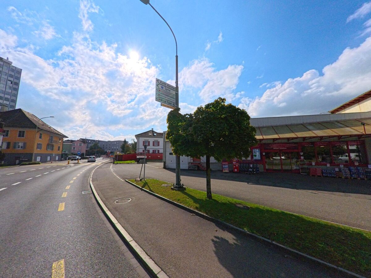 Eine breite Strasse in Burg (5737, Kanton Aargau) führt vorbei an einem Lebensmittelgeschäft mit rotem Eingangsbereich und Wohnhäusern unter einem klaren Himmel.