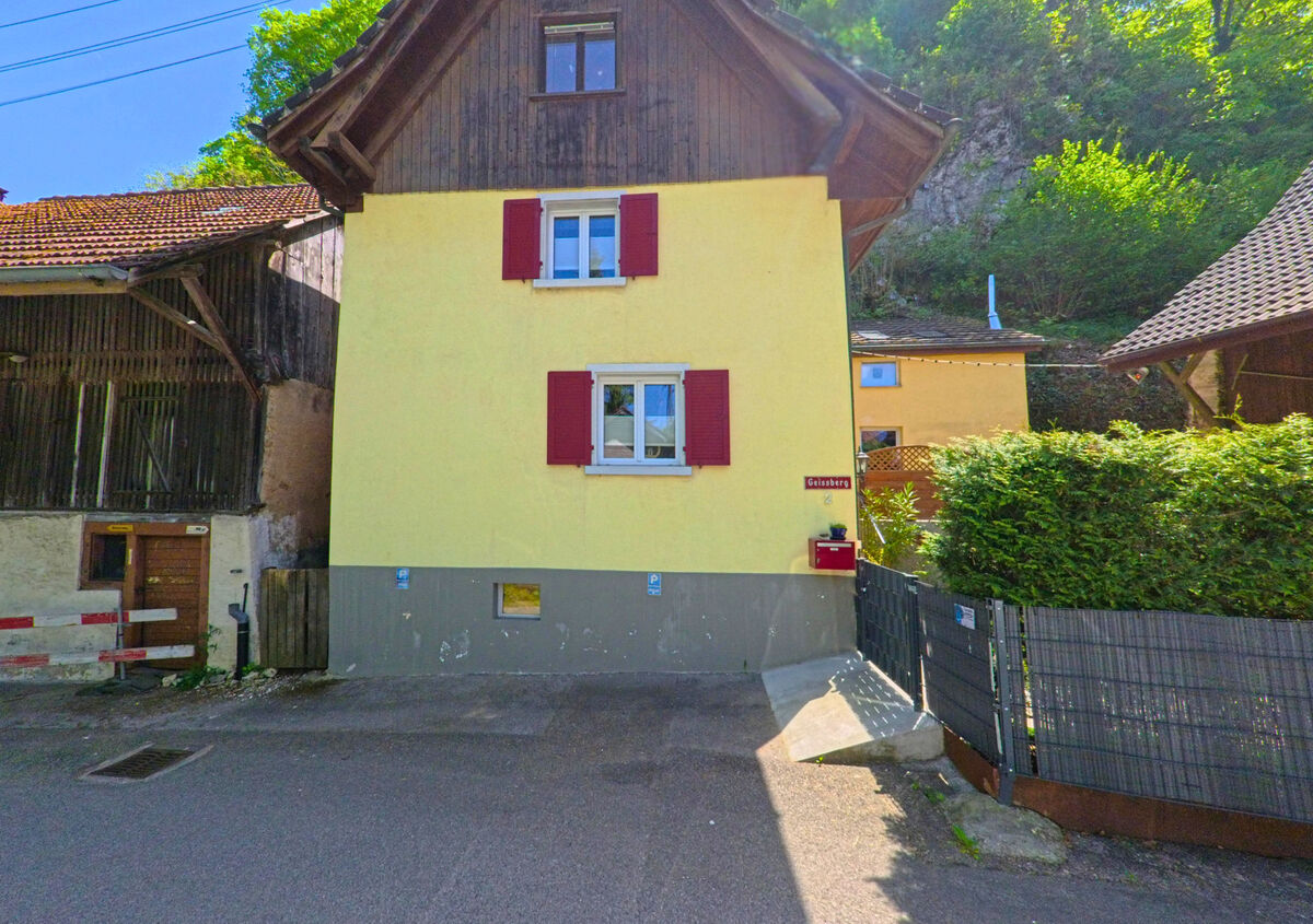 In Burg im Leimental (4117, Kanton Baselland) steht ein gelbes Wohnhaus mit roten Fensterlaeden an der Strasse „Geissberg“, umgeben von Scheune, Hang und Hecke.