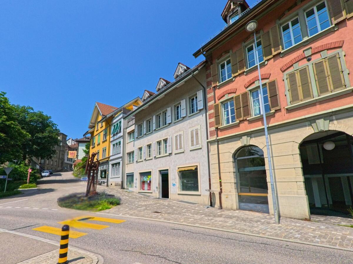 In der Altstadt von Burgdorf (3400, Kanton Bern) reihen sich farbenfrohe Fassaden entlang einer gepflasterten Strasse mit einer modernen Skulptur im Vordergrund.