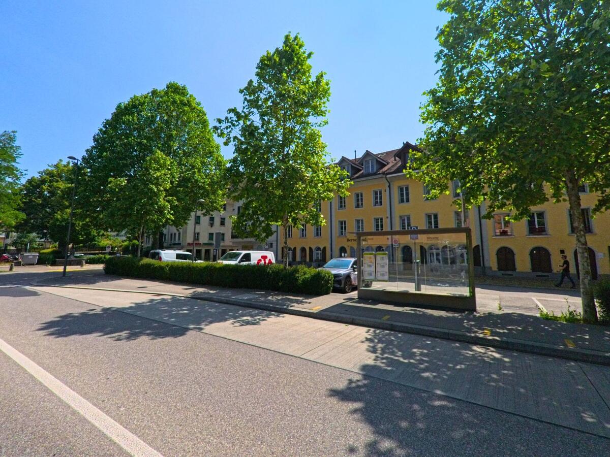 In Burgdorf (3401, Kanton Bern) befindet sich eine moderne Bushaltestelle vor einer Reihe gelber Altstadthäuser, eingerahmt von Schatten spendenden Bäumen.