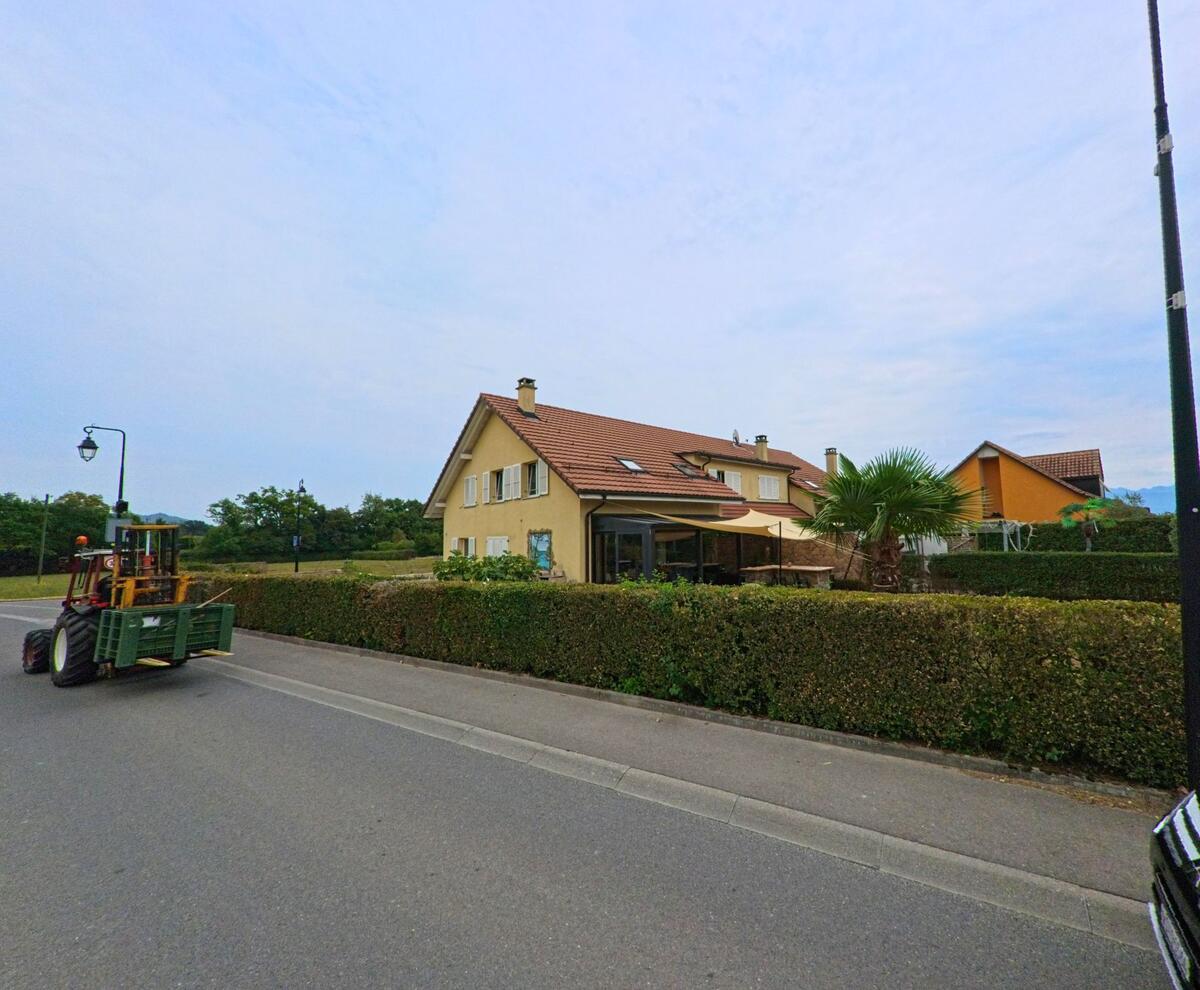 Ein Bauernfahrzeug faehrt an einem Einfamilienhaus mit Gartenhecke und Palmen in Bursinel (1195, Kanton Waadt) vorbei.