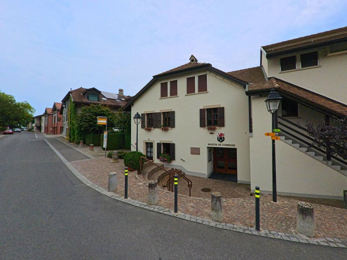 Das Gemeindehaus von Bursinel (1195, Kanton Waadt) mit heller Fassade und Gemeindewappen befindet sich an einer gepflasterten Strasse mit Bushaltestelle.