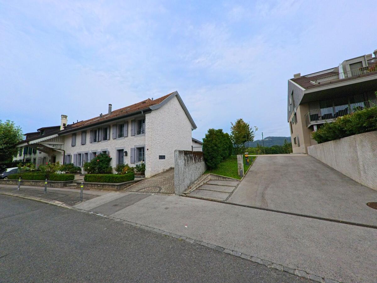 Ein traditionelles Wohnhaus mit hellen Fensterlaeden steht gegenueber einem modernen Mehrfamilienhaus in Bursinel (1195, Kanton Waadt).