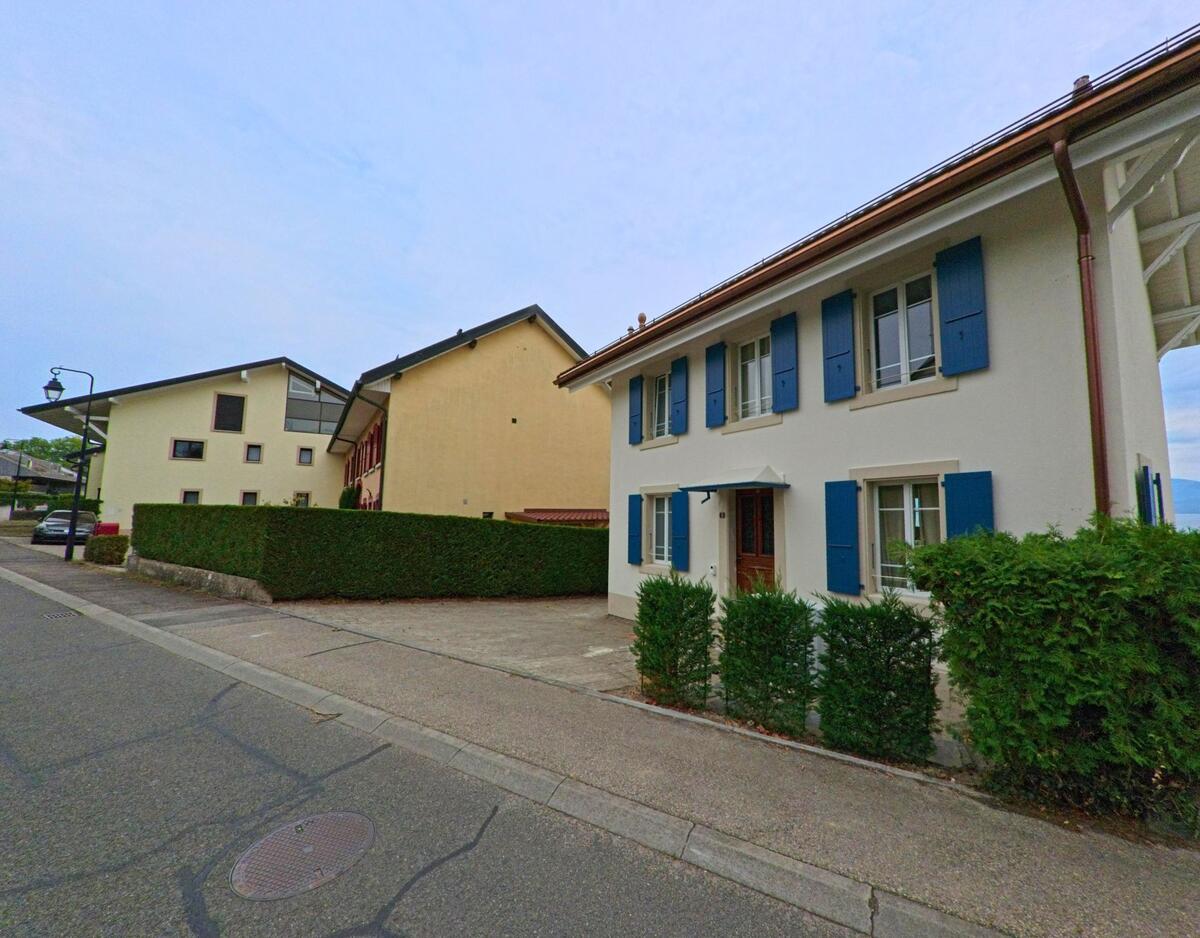 Ein weisses Wohnhaus mit blauen Fensterlaeden steht an einer ruhigen Strasse in Bursinel (1195, Kanton Waadt), daneben moderne Gebaeude mit gelben Fassaden.