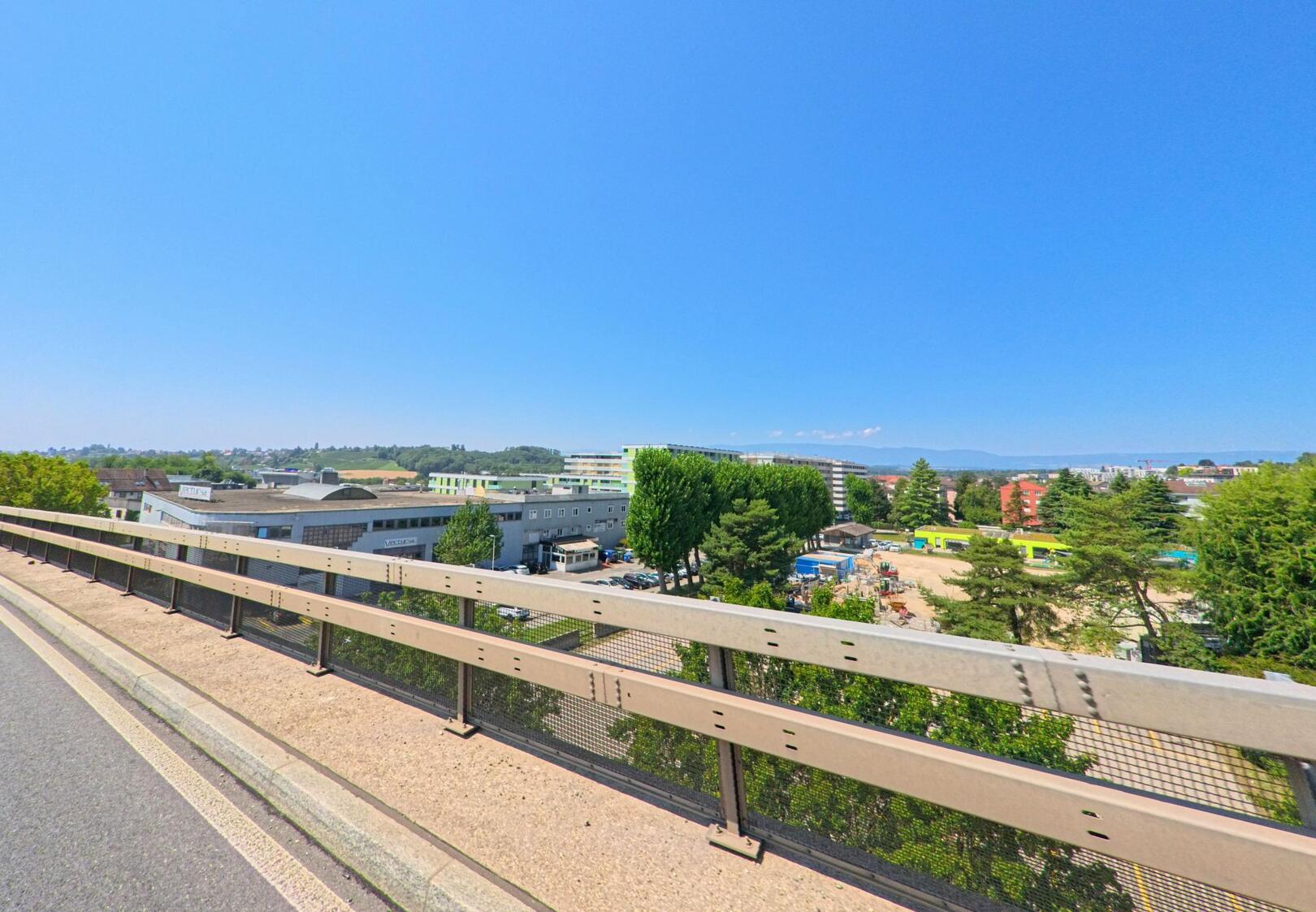 Von einer Brücke in Bussigny (1030, Kanton Waadt) eröffnet sich der Blick auf Industriegebäude, Baustellenflächen und Wohnhäuser. Im Hintergrund sind grüne Bäume und Hügel zu erkennen, die das Stadtbild ergänzen.