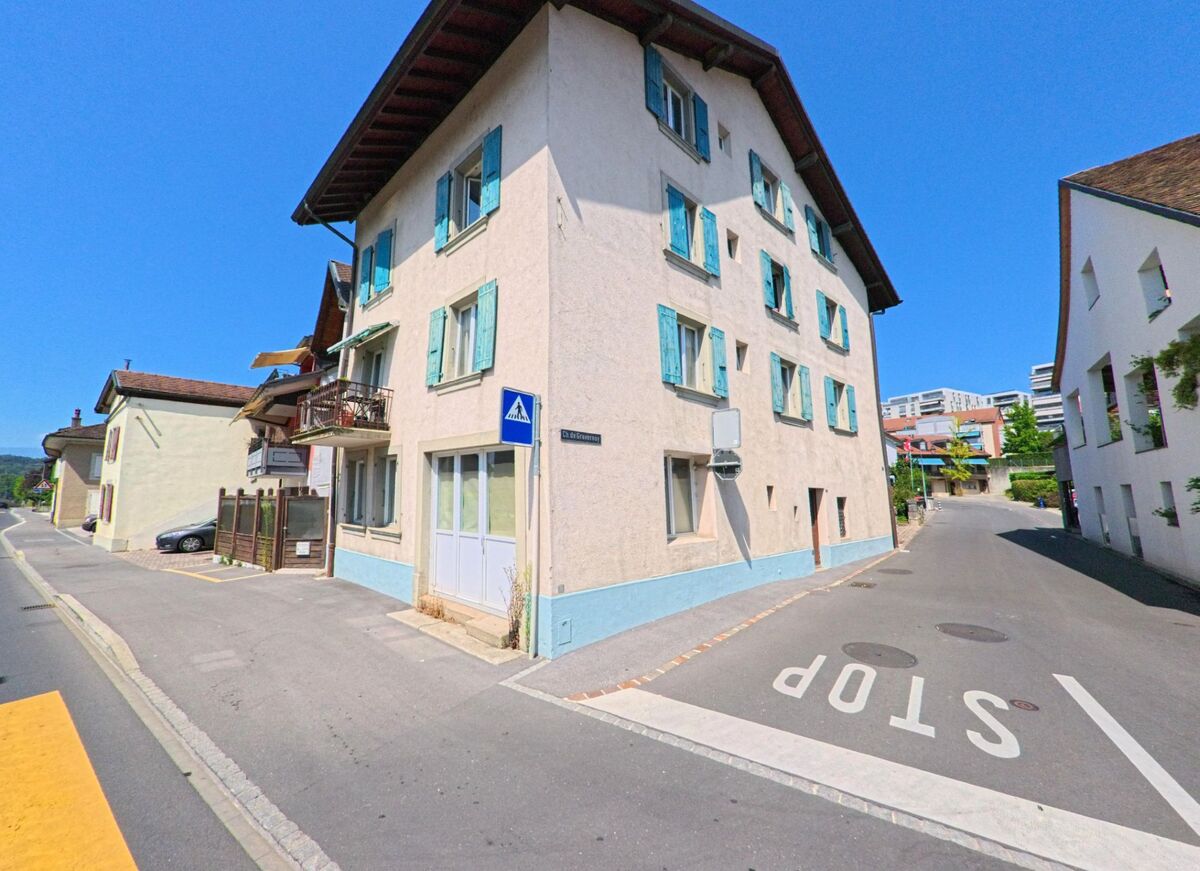 In Bussigny (1030, Kanton Waadt) stehen traditionelle Wohnhäuser mit farbigen Fensterläden an einer schmalen Strassengabelung. Ein Fussgängerstreifen und die Markierung „STOP“ strukturieren den ruhigen Strassenabschnitt.