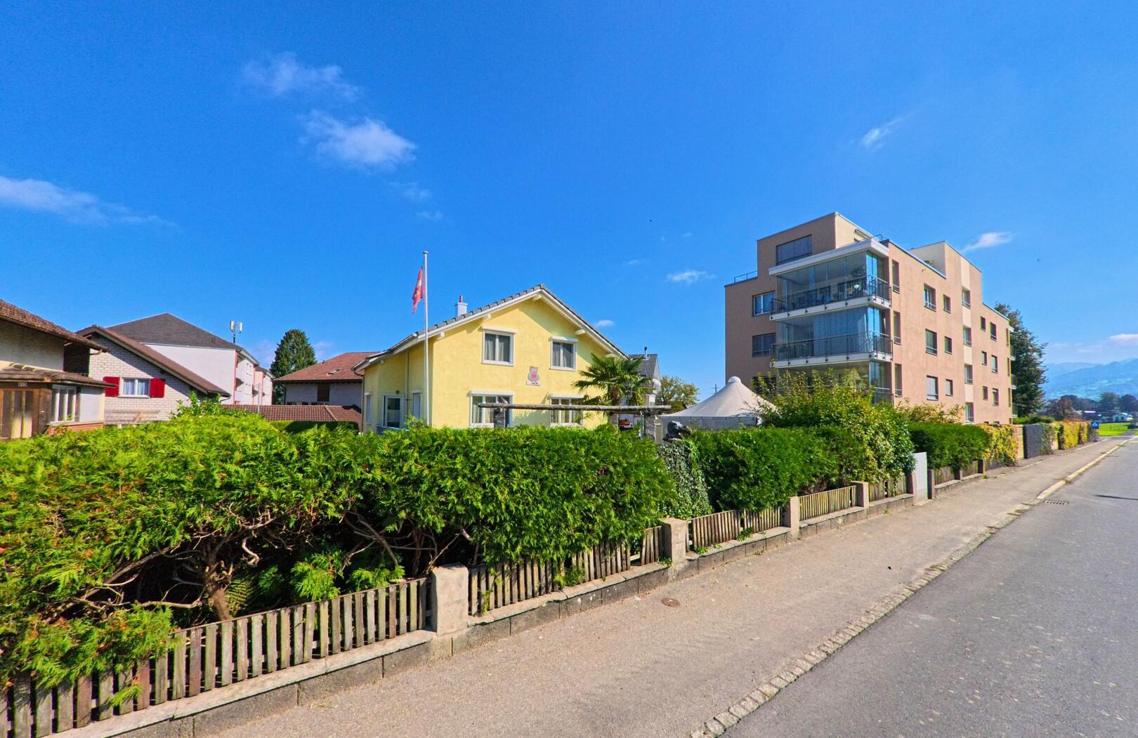 In Buttikon (8862, Kanton Aargau zeigt das Bild ein Wohnhaus mit gelber Fassade, das von einem hohen grünen Heckenzaun umgeben ist. Im Hintergrund ist ein modernes Mehrfamilienhaus sichtbar, das sich entlang der Straße erstreckt.