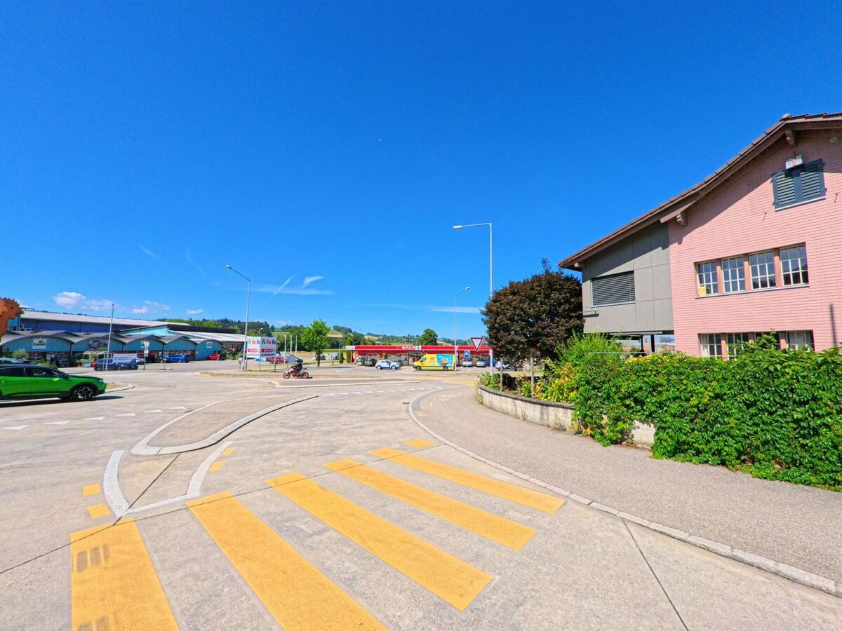 An einer grosszuegigen Einfahrt in Buttisholz (6018, Kanton Luzern) befinden sich mehrere Gewerbebauten sowie eine Tankstelle, daneben verlaeuft ein Fussgaengerstreifen unter klarem Himmel.